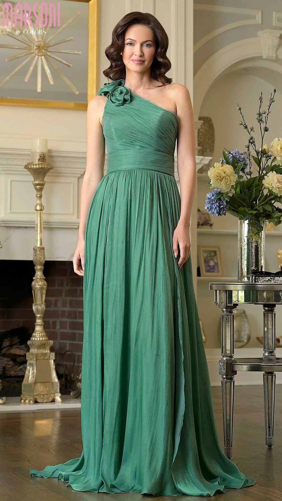 A woman poses gracefully by a fireplace and vase of flowers in the Marsoni M332 green long formal dress—a floor-length chiffon evening gown with an elegant one-shoulder design and striking 3D floral strap embellishment.